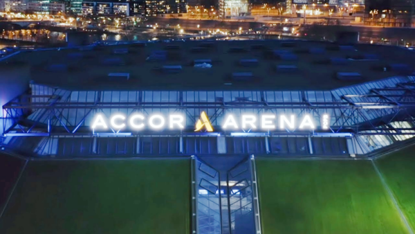 Vista aérea nocturna del exterior del Accor Arena con su letrero iluminado y las luces de la ciudad al fondo.