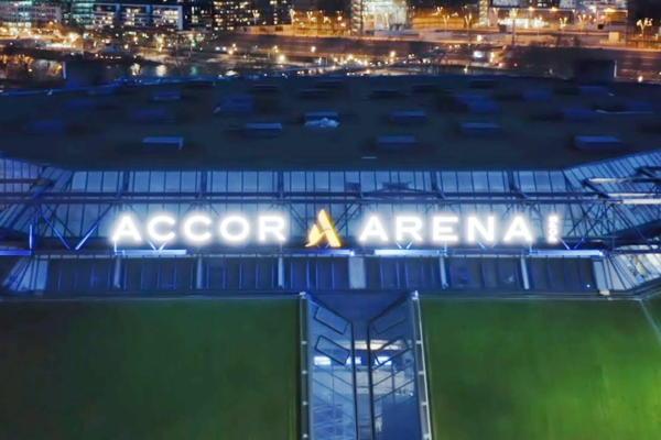 Vista aérea nocturna del exterior del Accor Arena con su letrero iluminado y las luces de la ciudad al fondo.