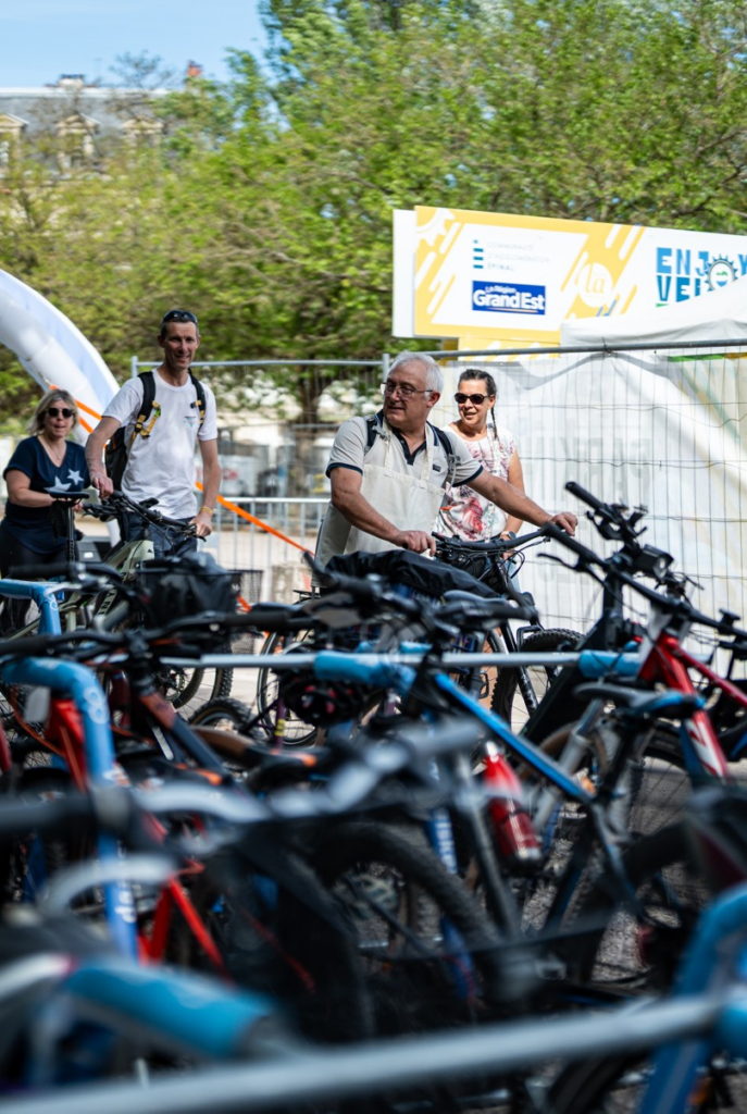 Grupo de personas con bicicletas en un evento ciclista al aire libre, en primer plano un hombre con chaleco beige y otros cerca con bicicletas detrás de una zona con pancartas.