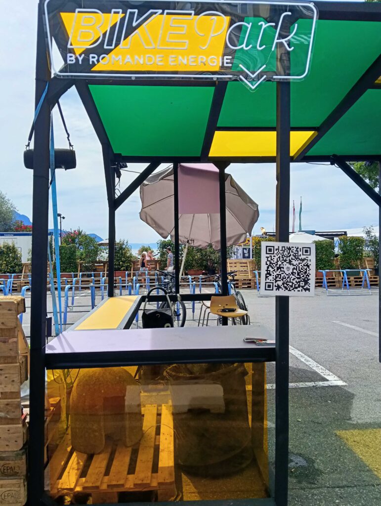 Quiosco de aparcamiento de bicicletas al aire libre con un letrero de neón 'BIKEpark', paneles verdes y amarillos, palés de madera y un letrero con código QR cercano; personas y bicicletas visibles al fondo.