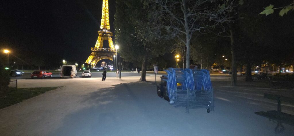 Vista nocturna de la Torre Eiffel iluminada en amarillo, vista desde un parque con sillas azules apiladas en primer plano.
